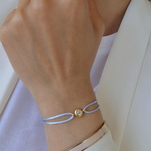 May include: A light blue bracelet with a gold accent and a clear crystal. The bracelet is tied with a knot and is worn on a person's wrist.
