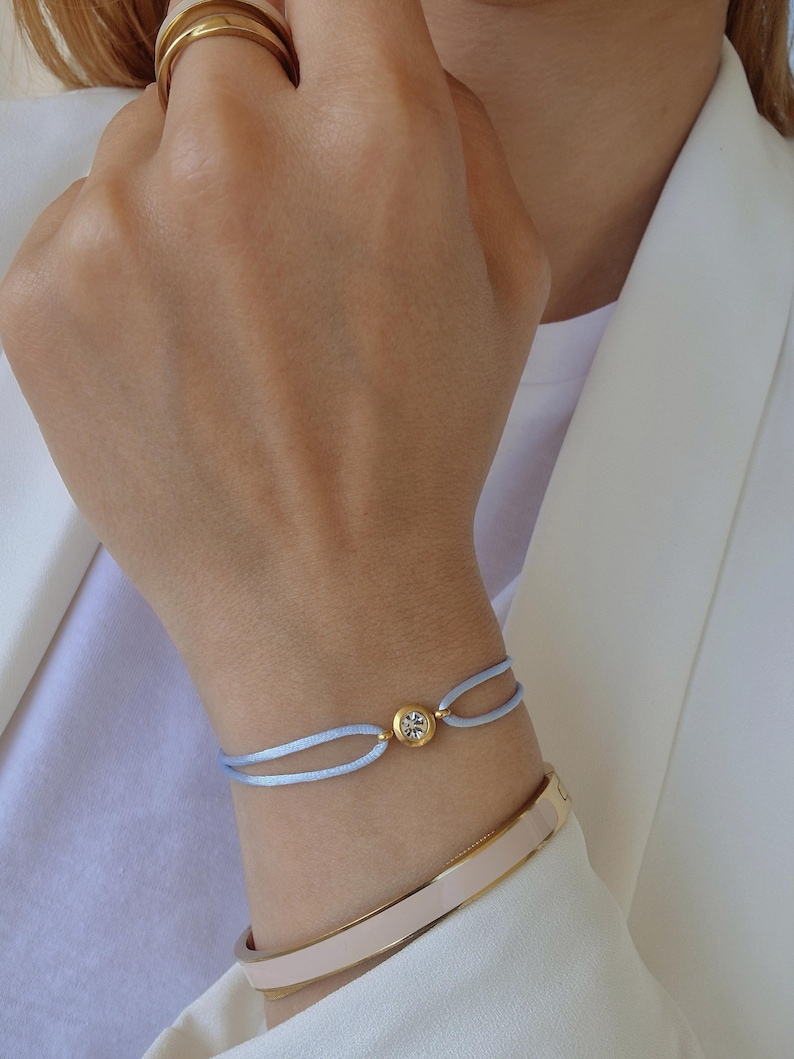 May include: A light blue bracelet with a gold accent and a clear crystal. The bracelet is tied with a knot and is worn on a person's wrist.