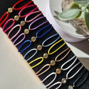 May include: A set of 12 colorful string bracelets with a single rhinestone charm on each. The bracelets are displayed on a black surface.