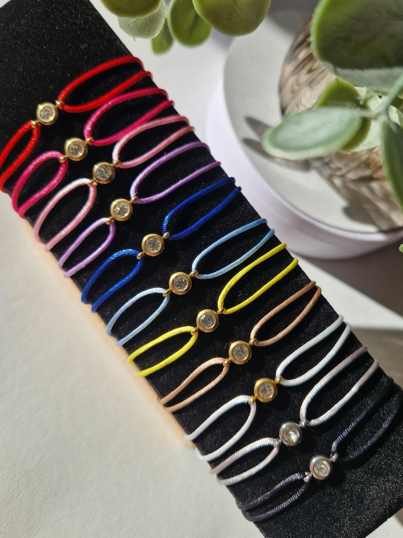 May include: A set of 12 colorful string bracelets with a single rhinestone charm on each. The bracelets are displayed on a black surface.