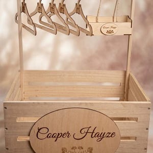 May include: A wooden clothing rack and storage crate set. The rack features butterfly-shaped hangers and a small shelf. The crate has an oval plaque with the name "Cooper Hayze" and floral accents. The set is made of light-colored wood.