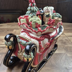 Puede incluir: Un trineo navideño de cerámica roja y blanca, adornado con regalos, un árbol de Navidad y una figura de ángel. Los patines del trineo son negros con detalles dorados, creando un ambiente festivo.