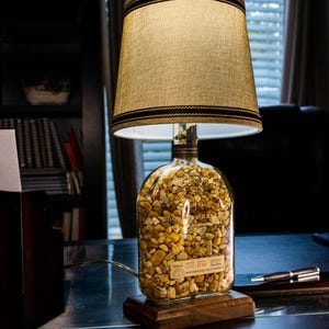 May include: A table lamp made from a clear glass bottle filled with small, light brown and yellow stones. The bottle has a label that reads "Woodford Reserve" and "750ml 10003 8740". The lamp has a brown fabric shade with a black trim.
