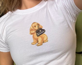Puede incluir: Camiseta blanca de cuello redondo con una ilustración en acuarela de un Cocker Spaniel dorado sosteniendo un bolso negro y blanco. El perro tiene pelaje marrón. La camiseta está hecha de un material suave y cómodo.