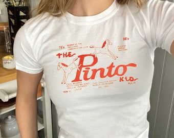 Może przedstawiać: Biała koszulka z czerwonym nadrukiem dwóch koni i tekstem "The Pinto Kid" z dodatkowym tekstem o restauracji.
