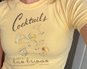 Może przedstawiać: Żółta koszulka z vintage'owym wzorem przedstawiającym cherubina trzymającego kieliszek koktajlowy i tekst "Cocktails Hotel las brisas Acapulco, Meksyk".