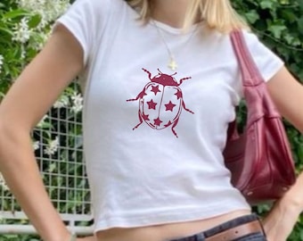May include: White t-shirt with a red and white ladybug graphic, paired with black denim jeans, a brown belt, and black flats. A burgundy handbag is carried over the shoulder. The person is wearing sunglasses.