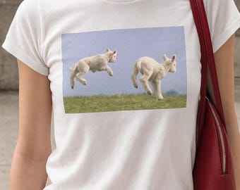 Peut inclure: T-shirt blanc avec un motif rectangulaire représentant deux agneaux blancs gambadant sur une colline herbeuse sous un ciel bleu clair. Les agneaux sont représentés en plein saut, créant une scène ludique. Le t-shirt est en matière blanche et douce.