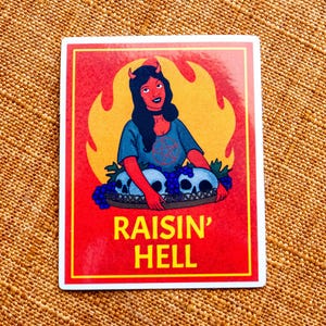 May include: A red sticker with a yellow flame design. A woman with red horns and a blue shirt is holding a basket of skulls. The text "raisin' hell" is printed in white letters.