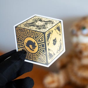 Op de afbeelding: Een sticker van een gouden kubus met zwarte kattenillustraties en een zwart katten silhouet aan één kant. De kubus is een gestileerde weergave van een puzzeldoos.