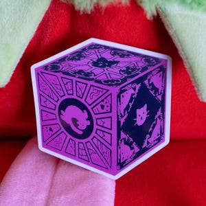 Op de afbeelding: Een paarse en witte sticker van een kubus met een kattenontwerp. De kubus heeft een zonnestralenpatroon en een kattengezicht aan elke kant.