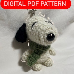 Pode incluir: Um chaveiro Snoopy de crochê com uma echarpe verde, orelhas pretas e um nariz preto. A imagem inclui o texto "DIGITAL PDF PATTERN" e "*NOT PHYSICAL SNOOPY KEYCHAIN*". O chaveiro é branco e tem um fecho de metal.