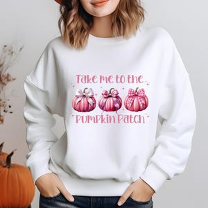 Peut inclure: Sweat-shirt blanc avec un motif rose de trois citrouilles avec des nœuds roses et le texte "Take me to the Pumpkin Patch".