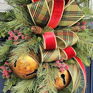 May include: A holiday door swag featuring green pine branches, two large rustic gold metal jingle bells, and clusters of frosted red berries. The arrangement is accented with a multi-loop bow made of red velvet ribbon and a tan, green, and black plaid ribbon.