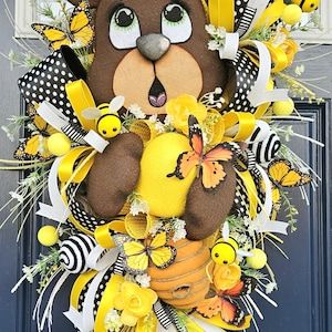 May include: A whimsical wreath featuring a brown bear with a surprised expression, surrounded by yellow and black accents. The wreath includes butterflies, bees, ribbons, and a honeycomb, creating a cheerful, nature-inspired design.