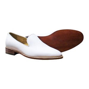 Op de afbeelding: Een paar witte leren loafers met een gladde afwerking, een lage gestapelde houten hak en een donkerbruine leren zool. Het instapmodel heeft een ronde neus en een beige voering, waarbij zowel het zijprofiel als de onderkant van de zool zichtbaar zijn.