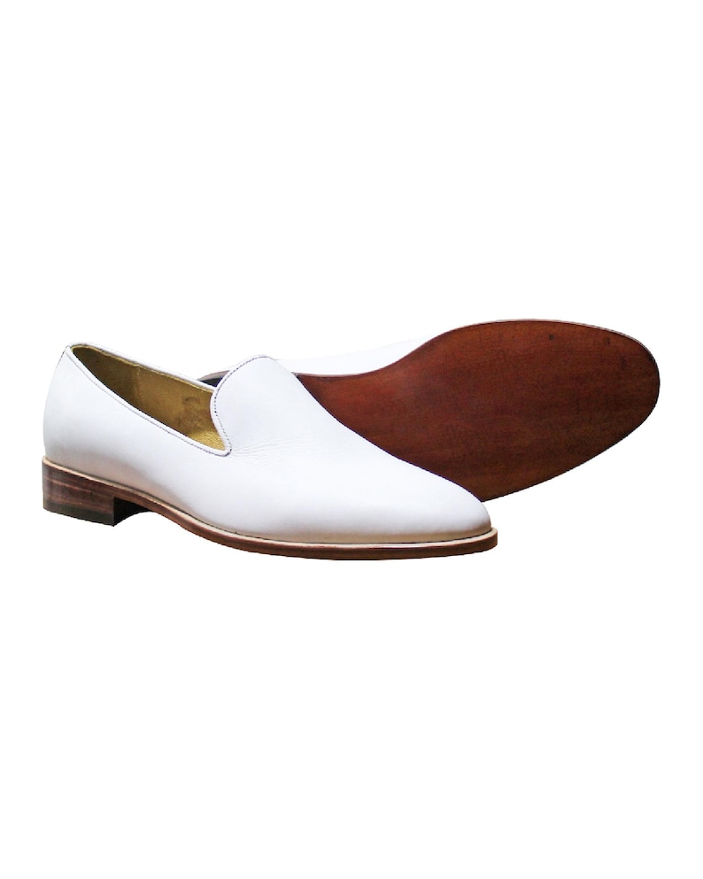 Op de afbeelding: Een paar witte leren loafers met een gladde afwerking, een lage gestapelde houten hak en een donkerbruine leren zool. Het instapmodel heeft een ronde neus en een beige voering, waarbij zowel het zijprofiel als de onderkant van de zool zichtbaar zijn.