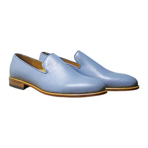 Op de afbeelding: Een paar lichtblauwe leren loafers met een gladde afwerking, een ronde neus en een laag profiel. De schoenen zijn voorzien van een beige leren tussenzool en een donkerbruine gelaagde hak, wat zorgt voor een strak en eenvoudig uiterlijk.