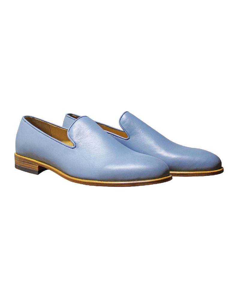 Op de afbeelding: Een paar lichtblauwe leren loafers met een gladde afwerking, een ronde neus en een laag profiel. De schoenen zijn voorzien van een beige leren tussenzool en een donkerbruine gelaagde hak, wat zorgt voor een strak en eenvoudig uiterlijk.