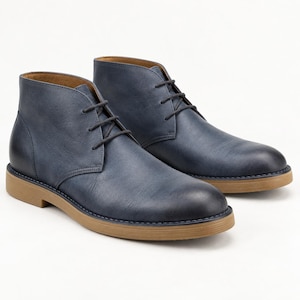 Könnte beinhalten: Ein Paar marineblaue Stiefeletten mit glatter Oberfläche, dunklen Schnürsenkeln und einer beigen Gummisohle. Die Stiefel haben eine abgerundete Zehenpartie und eine mittelhohe Silhouette mit sichtbaren Nähten entlang der Sohlenkante.