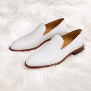 Op de afbeelding: Een paar witte leren instappers met een gladde afwerking, een ronde neus en een laag profiel. De schoenen zijn voorzien van een beige lederen voering en een houten hak met een donkerbruine rand langs de zool.