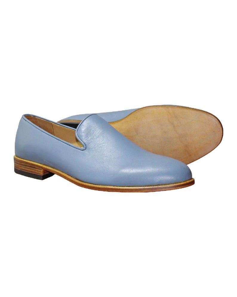 Op de afbeelding: Een paar lichtblauwe leren loafers met een gladde afwerking, een laag profiel, een beige gestapelde houten hak en een lichtbruine leren zool. De schoenen hebben een eenvoudig instapmodel met een ronde neus en een strak, onversierd bovenwerk.