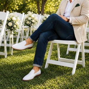 Op de afbeelding: Een paar witte leren instappers met een lage hak en ronde neus, gedragen met een donkerblauwe spijkerbroek, een wit overhemd en een beige blazer. De schoenen hebben een gladde afwerking en een bruine rand langs de zool.