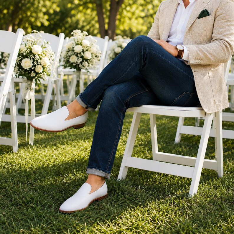 Op de afbeelding: Een paar witte leren instappers met een lage hak en ronde neus, gedragen met een donkerblauwe spijkerbroek, een wit overhemd en een beige blazer. De schoenen hebben een gladde afwerking en een bruine rand langs de zool.