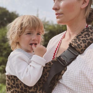 Könnte beinhalten: Eine Babytrage mit Leopardenmuster und schwarzen Riemen und Schnalle. Ein Kind in einem weißen Langarm-Oberteil befindet sich in der Trage. Die Person, die die Trage trägt, trägt ein weißes Oberteil mit roter Bordüre.