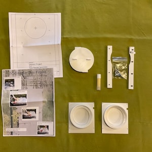 May include: A disassembled bee feeder kit, featuring white plastic components, screws, a small tube, and printed instructions. The instructions include the title "Sliding Jar Bee Feeder" and diagrams. The kit is arranged on a green surface.