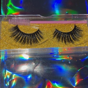Op de afbeelding: Een paar zwarte valse wimpers in een doorzichtige plastic doos met een gouden glitterbasis. Een roze wimperborstel is inbegrepen. De verpakking heeft een holografisch iriserend effect.