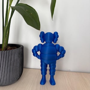 Peut inclure: Figurine bleue représentant un personnage de dessin animé avec des yeux barrés et un corps arrondi. La figurine a un design unique avec des membres arrondis et une forme de tête distinctive. Elle est posée sur une surface en bois, avec une plante en pot en arrière-plan.