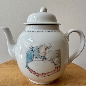 May include: White ceramic teapot featuring a Peter Rabbit illustration. The teapot depicts a scene of Peter Rabbit giving a cup of tea to a bunny in bed. The text on the teapot reads "...and she gave a dose of it to Peter".