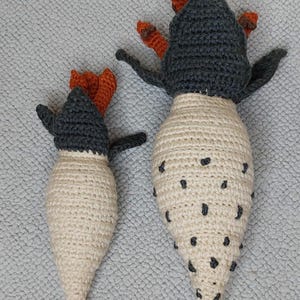 Könnte beinhalten: Zwei handgefertigte Häkelfiguren, die an Vögel oder Kreaturen erinnern. Die Figuren sind hauptsächlich cremefarben mit dunkelgrauen Köpfen und Flügeln. Sie haben orangefarbene Akzente auf ihren Köpfen und schwarze Punkte auf ihren Körpern. Eine ist größer als die andere.