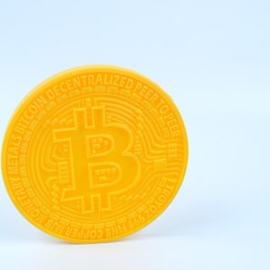 Pode incluir: Uma réplica de plástico amarela de uma moeda Bitcoin com o texto "2009 FINE COPPER 1 TROY OZ DIGITAL METALS BITCOIN DECENTRALIZED PEER TO PEER"