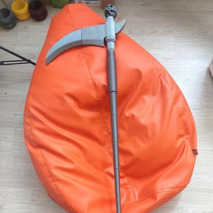 Könnte beinhalten: Eine graue und schwarze, teleskopierbare Stangenwaffe mit einer gebogenen Klinge und einem schädelförmigen Knauf. Die Waffe liegt auf einem leuchtend orangefarbenen Sitzsack.