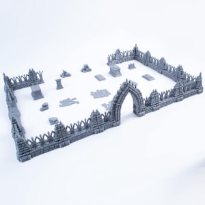 Op de afbeelding: Een grijze miniatuur begraafplaatsset met een stenen omheining met spitsbogen, een centrale boogpoort en diverse verspreide grafmarkeringen, grafstenen en stenen platen die binnen het rechthoekige gebied zijn gerangschikt.