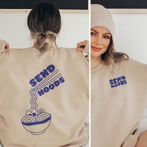 Op de afbeelding: Een beige sweatshirt met een blauwe afbeelding van eetstokjes die noedels in een kom vasthouden. De tekst "SEND NOODS" is geschreven in een retro-lettertype.
