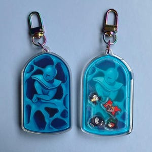 Peut inclure: Deux porte-clés en acrylique transparent avec fermoirs irisés. Chaque porte-clés présente une illustration bleue dans un cadre en forme d'arche. L'un des porte-clés présente une figure bleue, l'autre la même figure avec de petits breloques de personnages.