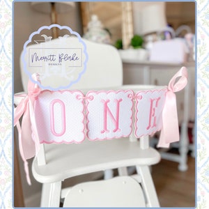 Può includere: Uno striscione rosa e bianco per seggiolone con la parola "ONE" ricamata in rosa. Lo striscione è decorato con pois bianchi e bordi a festoni. Lo striscione è legato con un nastro di raso rosa.