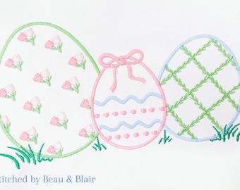 May include: Three embroidered Easter eggs in pink, blue, and green. The eggs are decorated with different patterns, including flowers, waves, and a grid. The eggs are sitting on a white background with green grass.