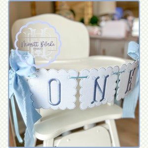 May include: A blue and white gingham banner with the word "ONE" embroidered in blue. The banner is strung across a white high chair with a blue ribbon tied to the back.
