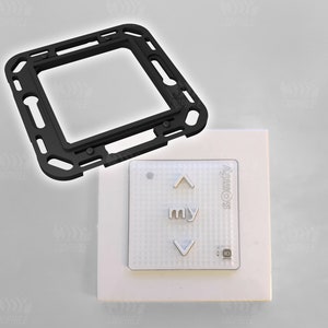 May include: A white square wall plate with a black frame. The plate has a white button with the word "my" and up and down arrows. The text "Somfy" is printed on the plate. A black plastic frame is shown separately.