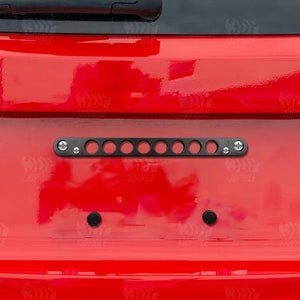May include: Black number plate frame with a row of round holes. The frame is attached to a red car.