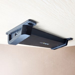 Op de afbeelding: Een zwart apparaat met een USB-poort is met een zwarte plastic beugel onder een houten oppervlak gemonteerd. Het apparaat heeft de tekst "Synology" erop gedrukt.