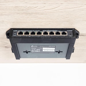 Puede incluir: Un switch inteligente Gigabit de 8 puertos negro con un soporte impreso en 3D. El switch tiene múltiples puertos y está montado sobre una superficie de madera clara. La marca "tp-link" es visible.