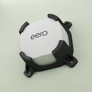 May include: A black plastic wall mount for an eero wifi router. The mount has a square base and four prongs that extend out to hold the router in place. The router is white and has the word "eero" printed on it.