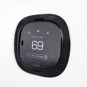Puede incluir: Un termostato inteligente Ecobee negro con una pantalla digital que muestra una temperatura de 69 grados Fahrenheit. El termostato está montado en una pared blanca.