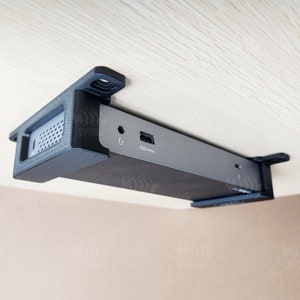 May include: A black and silver device with a USB port and a power port is mounted under a wooden surface. The device is held in place by a black plastic bracket.