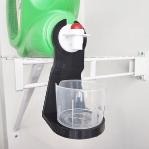 Può includere: Un erogatore in plastica nera con un pulsante rosso e una tazza graduata trasparente fissata a uno scaffale in metallo bianco. L'erogatore è progettato per contenere una grande bottiglia di liquido, come detersivo o soluzione detergente.